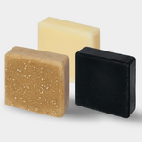 A trio of tallow soap bars arranged from front to back, each varying in color from a creamy yellow to a soft beige and finally to a deep slate blue, indicating different natural ingredients and scents. Each bar showcases a tactile, textured surface, indicative of their handcrafted nature, standing against a clean, neutral background that highlights their simple, yet elegant aesthetic.