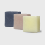 A trio of tallow soap bars arranged from front to back, each varying in color from a creamy yellow to a soft beige and finally to a deep slate blue, indicating different natural ingredients and scents. Each bar showcases a tactile, textured surface, indicative of their handcrafted nature, standing against a clean, neutral background that highlights their simple, yet elegant aesthetic.