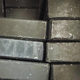 A close-up view of stacked charcoal facial soap bars arranged in an interlocking pattern. The bars have an organic, hand-cut shape and a deep grey color marbled with lighter streaks and flecks. The textured surface of the soap bars is matte and dusted with fine white particles, reminiscent of natural mineral deposits, giving a raw and earthy appeal to the product.