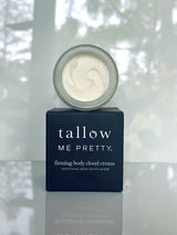 Tallow skin care Firming Body Cloud Cream in a jar with a creamy white texture, placed on a black box, set against a greenery and tile reflection backdrop.