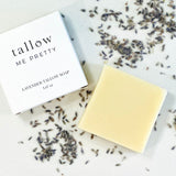 An elegant square bar of Lavender Tallow Soap lies next to its packaging, which is a clean, white box with 'Tallow Me Pretty' branding. The soap and box are set against a textured background scattered with dried lavender buds.