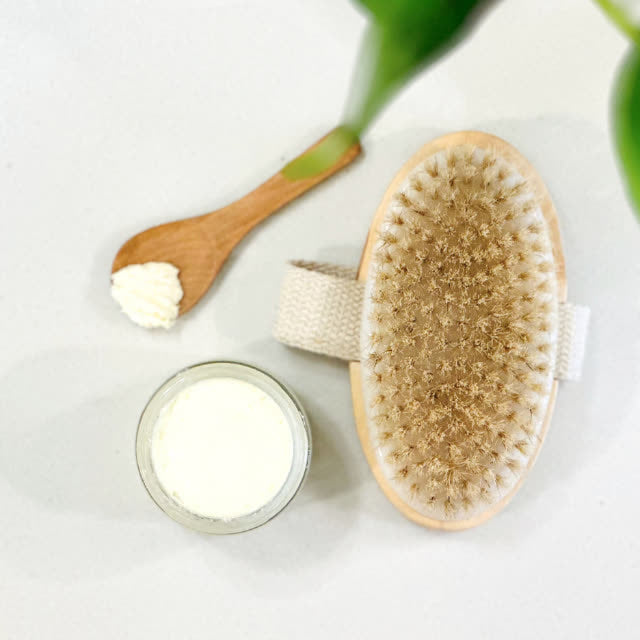 Overhead view of a natural bristle dry brush, wooden spoon with tallow balm, and open jar, accompanied by a fresh green plant leaf, representing a holistic approach to skin wellness.